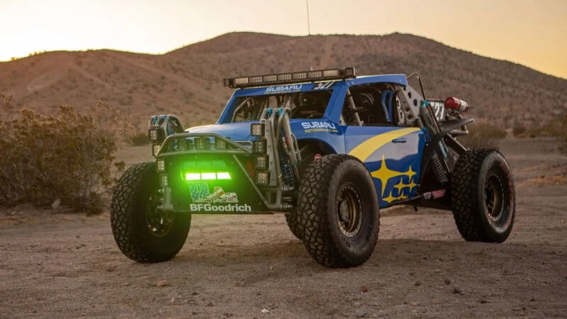 Blue Subaru Crosstrek Desert Racer with green BFGoodrich tires parked in sandy terrain with desert hills at sunset