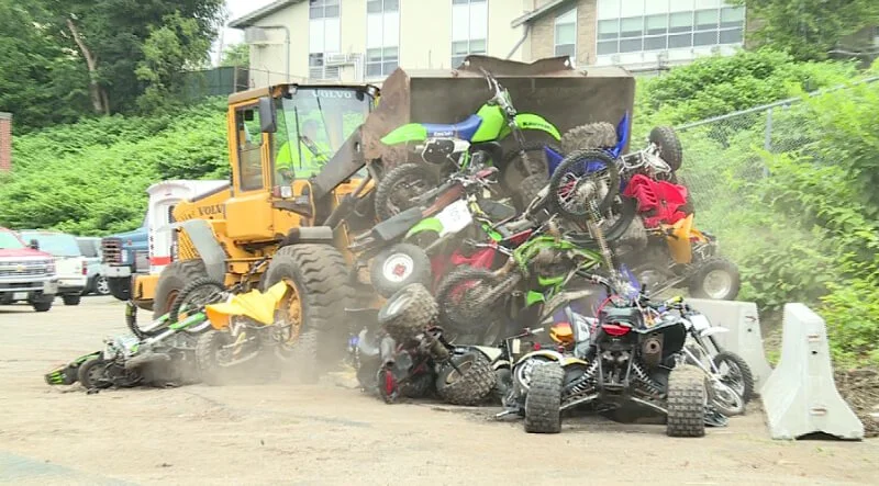 Yellow wheel loader crushing confiscated dirt bikes and ATVs in pile at public destruction event