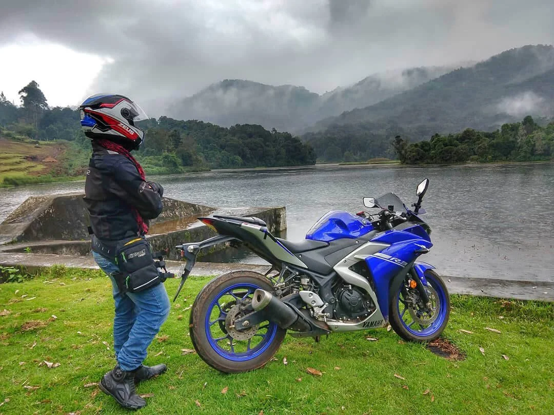 Motorcyclist in safety gear standing beside blue sport bike by scenic riverside lake surrounded by misty mountains