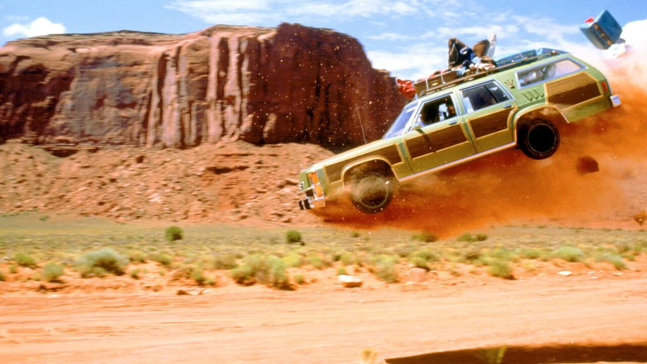 Vintage station wagon jumping through desert dust with rock formation backdrop