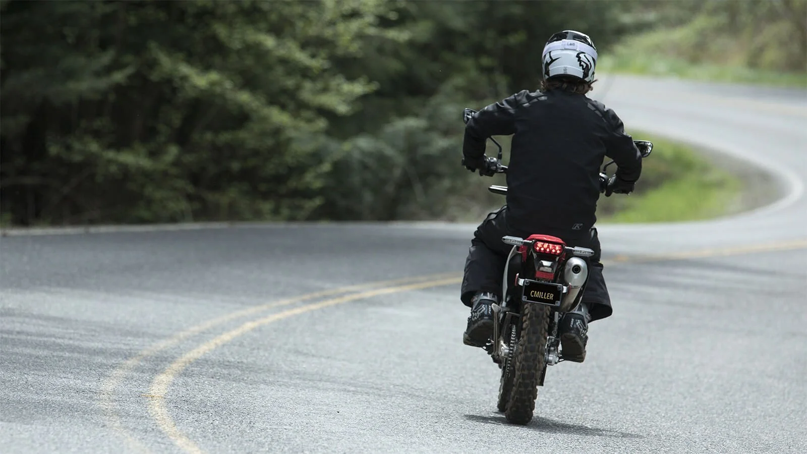 Rider on street legal dirt bike riding on curved asphalt road with forest background and yellow center line markings