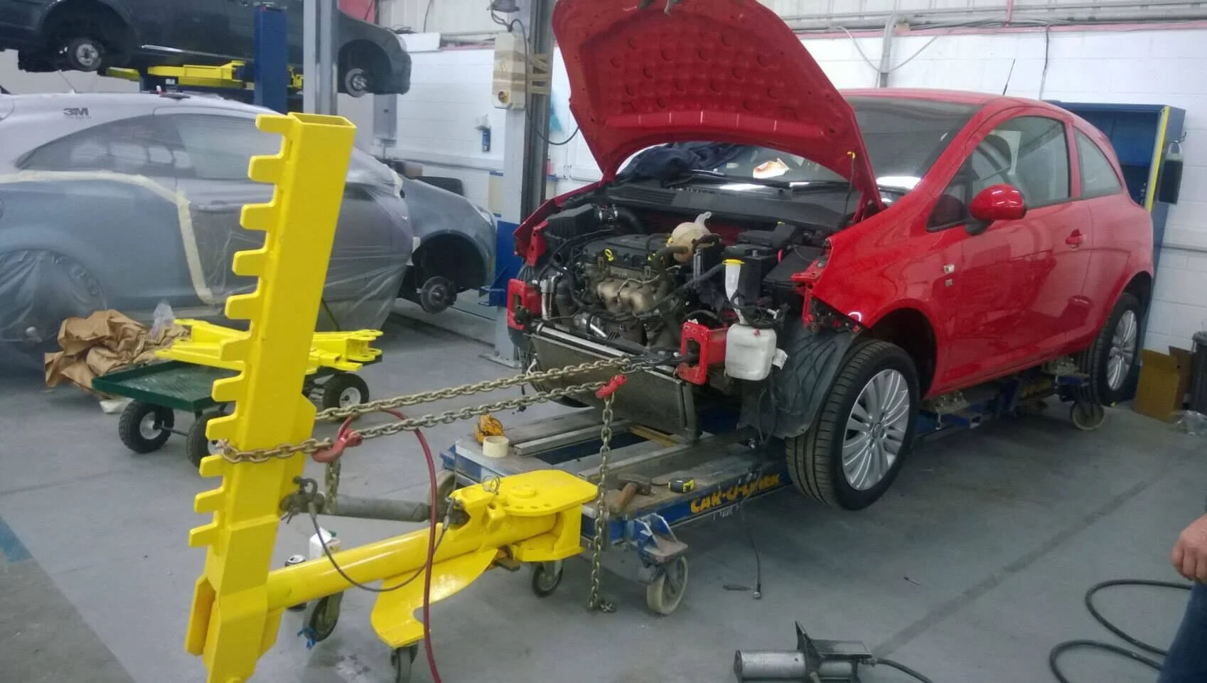 Red compact car undergoing frame straightening repair at auto body shop using yellow alignment equipment