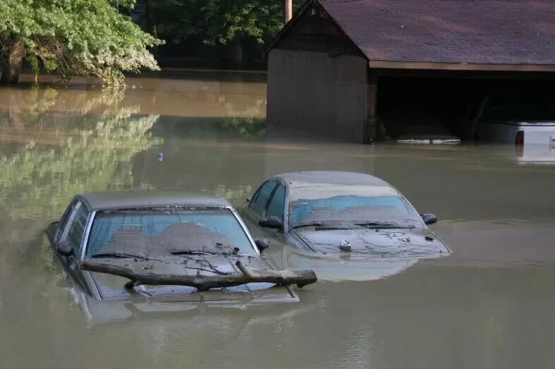 Two cars submerged in floodwater with buildings and trees in background, illustrating flood damage risks to vehicles