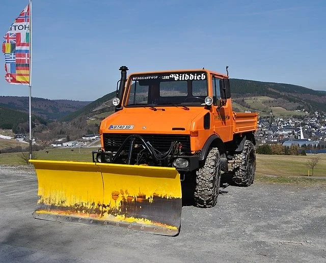 Orange Mercedes-Benz Unimog truck with yellow snow plow attachment in alpine landscape