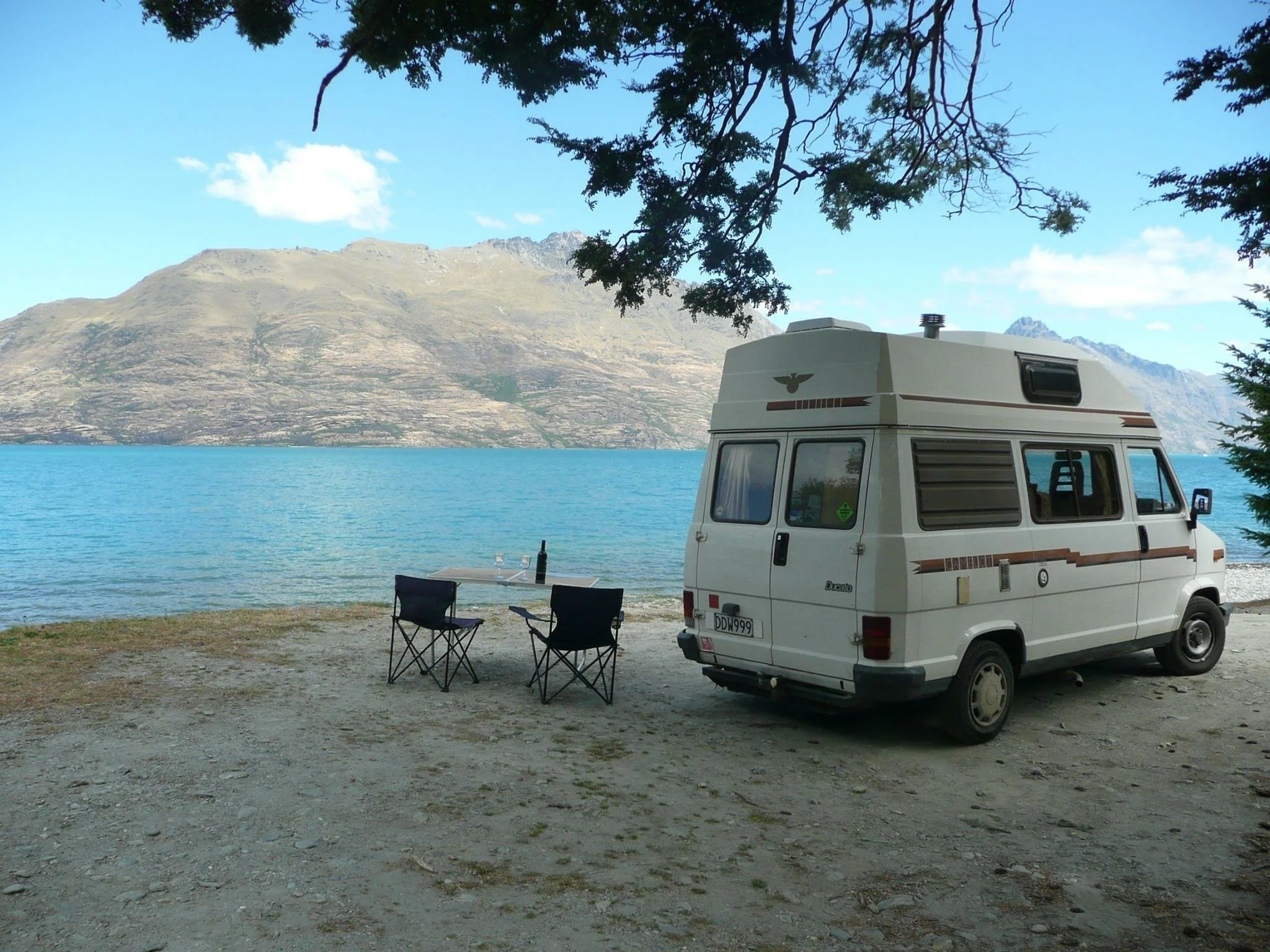 White camper van parked on beach with mountain lake view, camping chairs, and wine bottle for van life adventure
