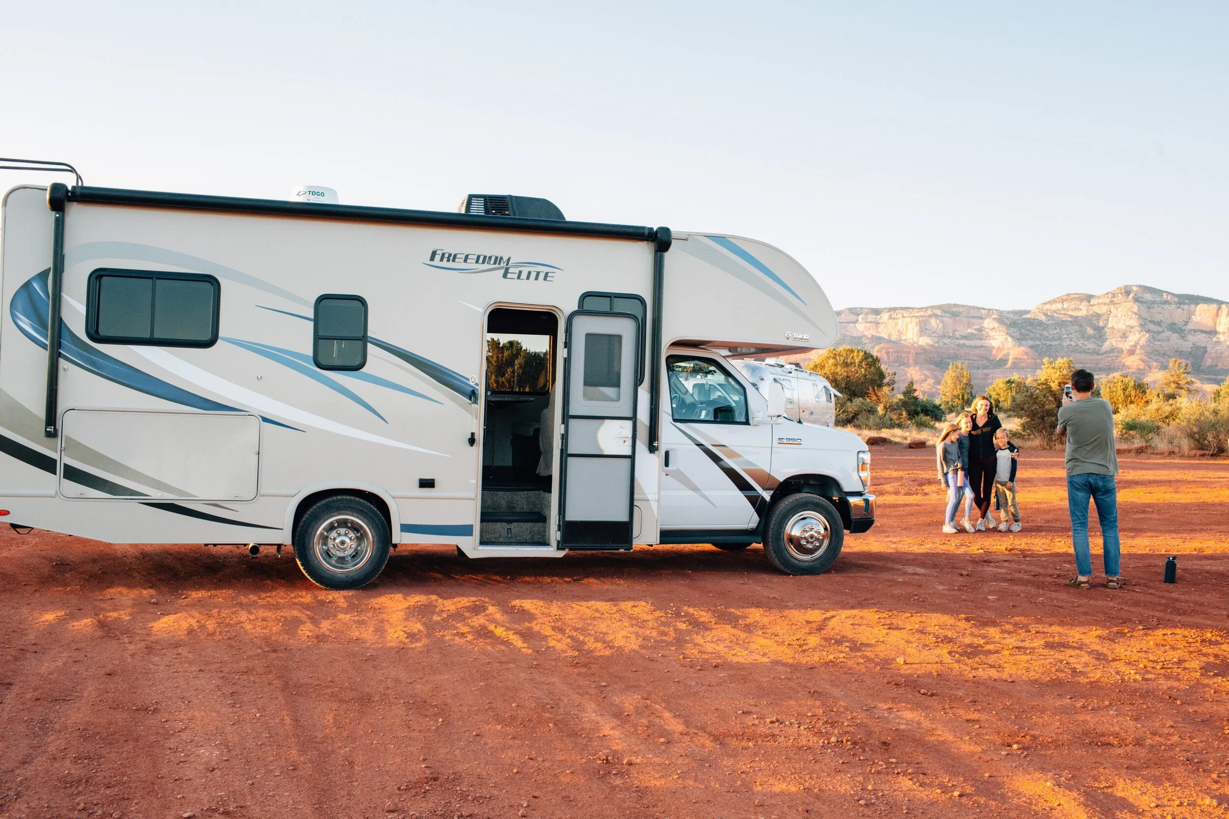 Family camping with white Freedom Elite RV in desert landscape with red rock mountains backdrop