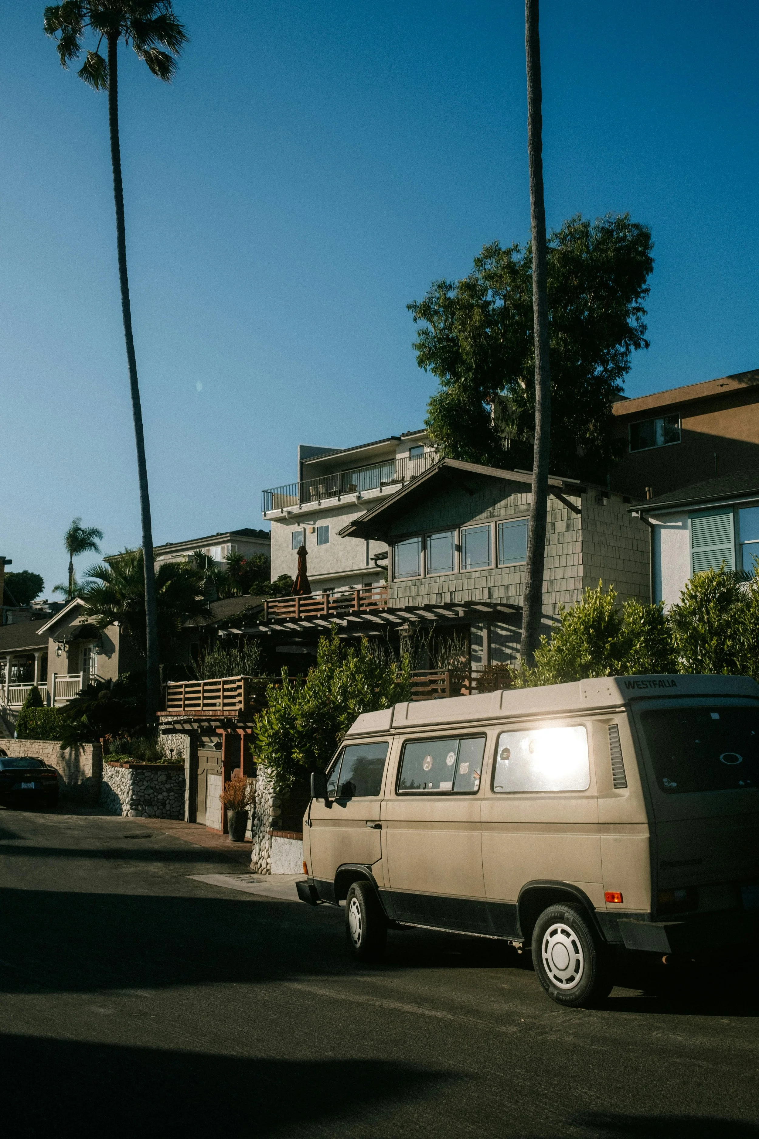 Beige van parked on residential street with modern house and tall palms