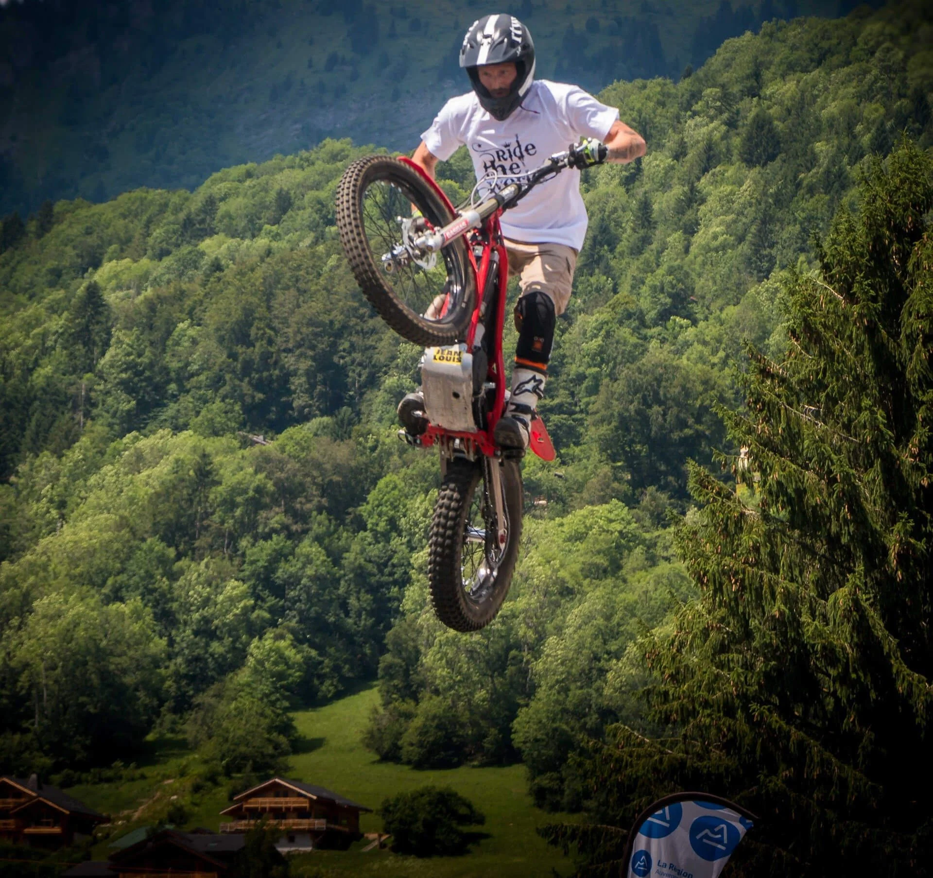 Professional rider performing aerial trials bike stunt over forested Alpine valley landscape