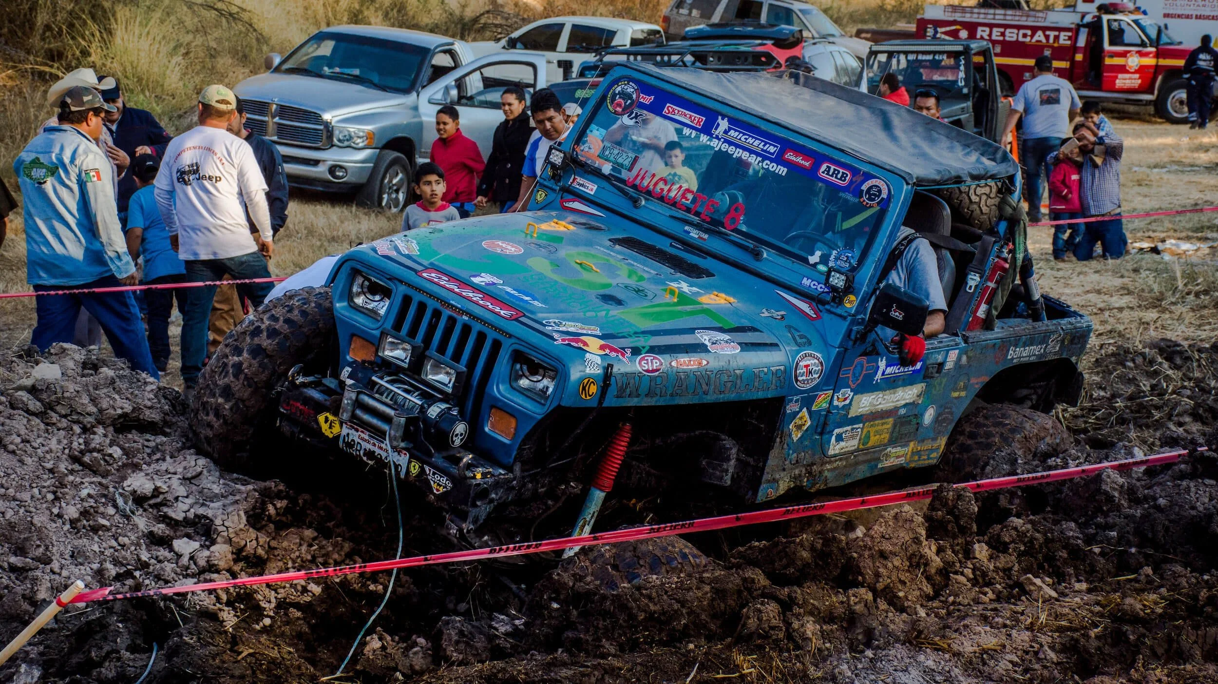 Blue Jeep Wrangler rock crawling through muddy terrain at off-road competition event with spectators watching