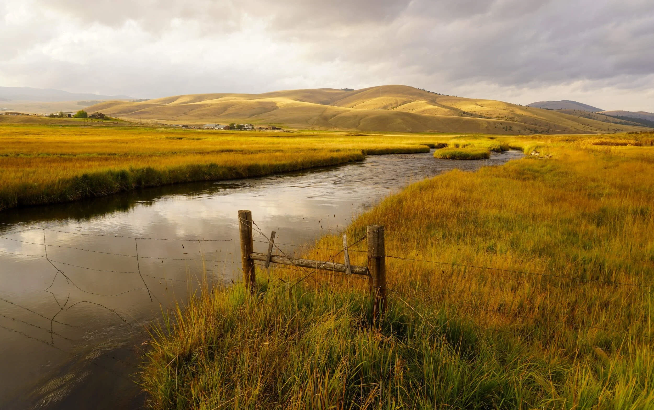 Rural Montana landscape with golden grassland, stream, and rolling hills under cloudy sky
