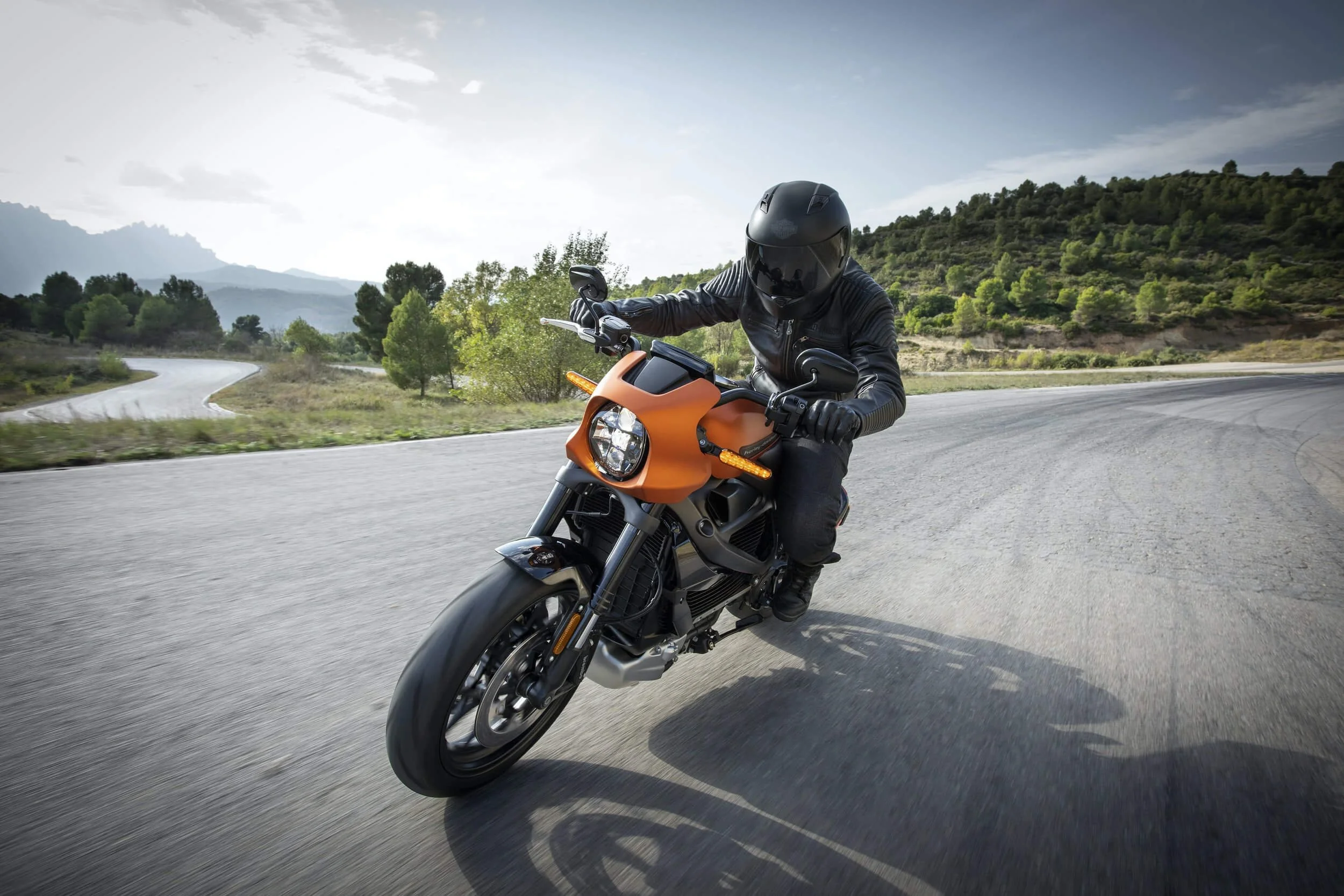 Rider on orange electric motorcycle leaning into turn on scenic mountain road with river landscape