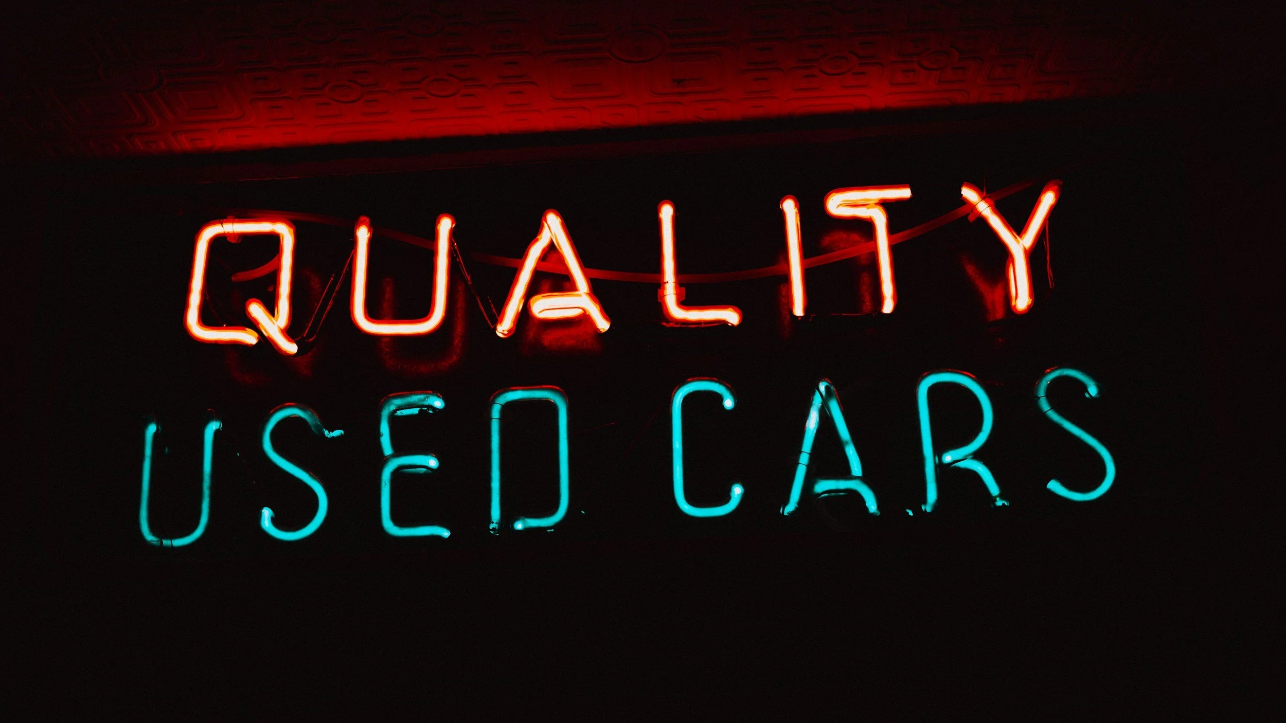 Red and cyan neon sign reading "Quality Used Cars" against dark background for vehicle sales business