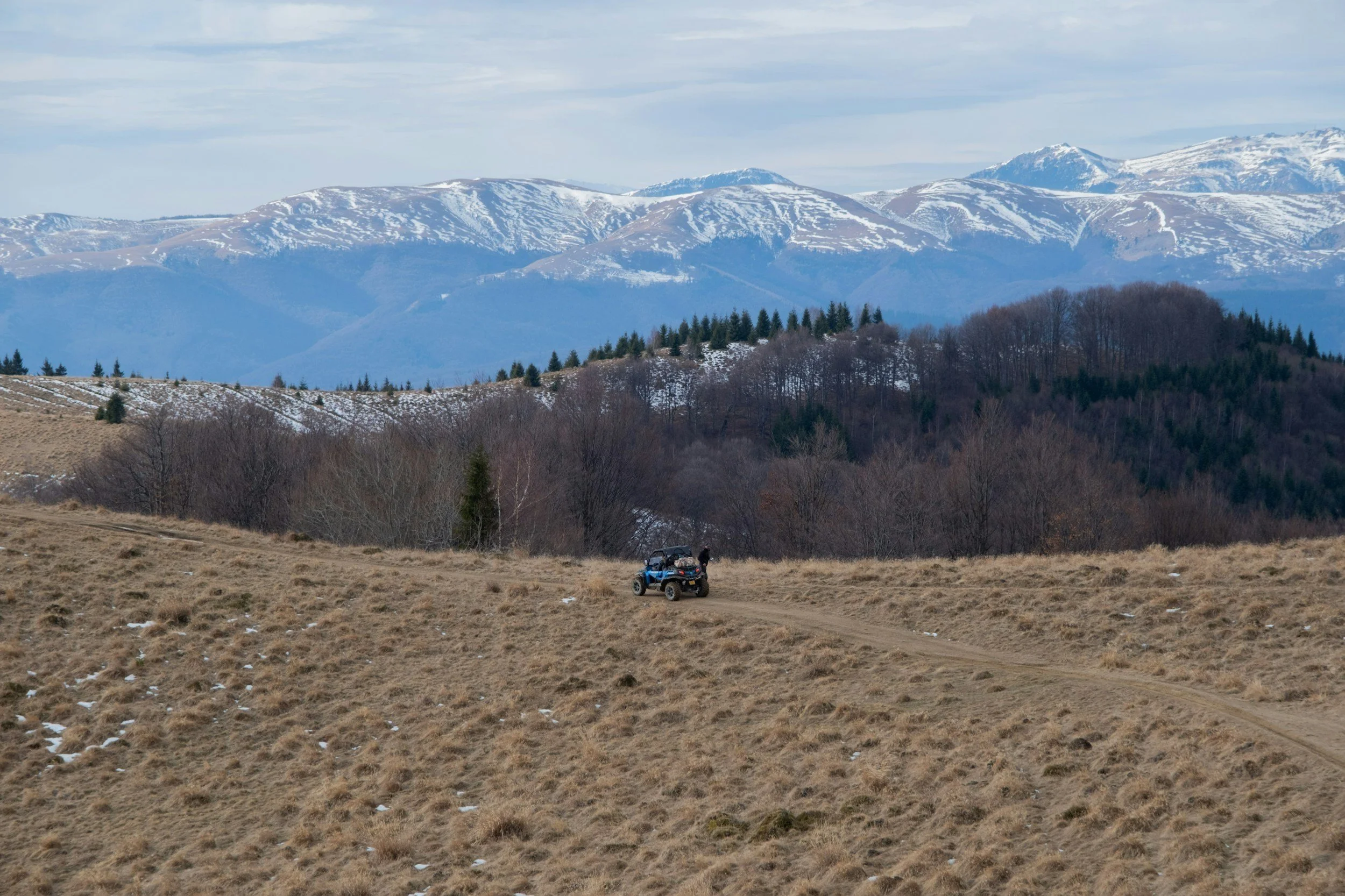 Blue ATV driving on dirt road through high mountain valley with snow-capped peaks and forest in background