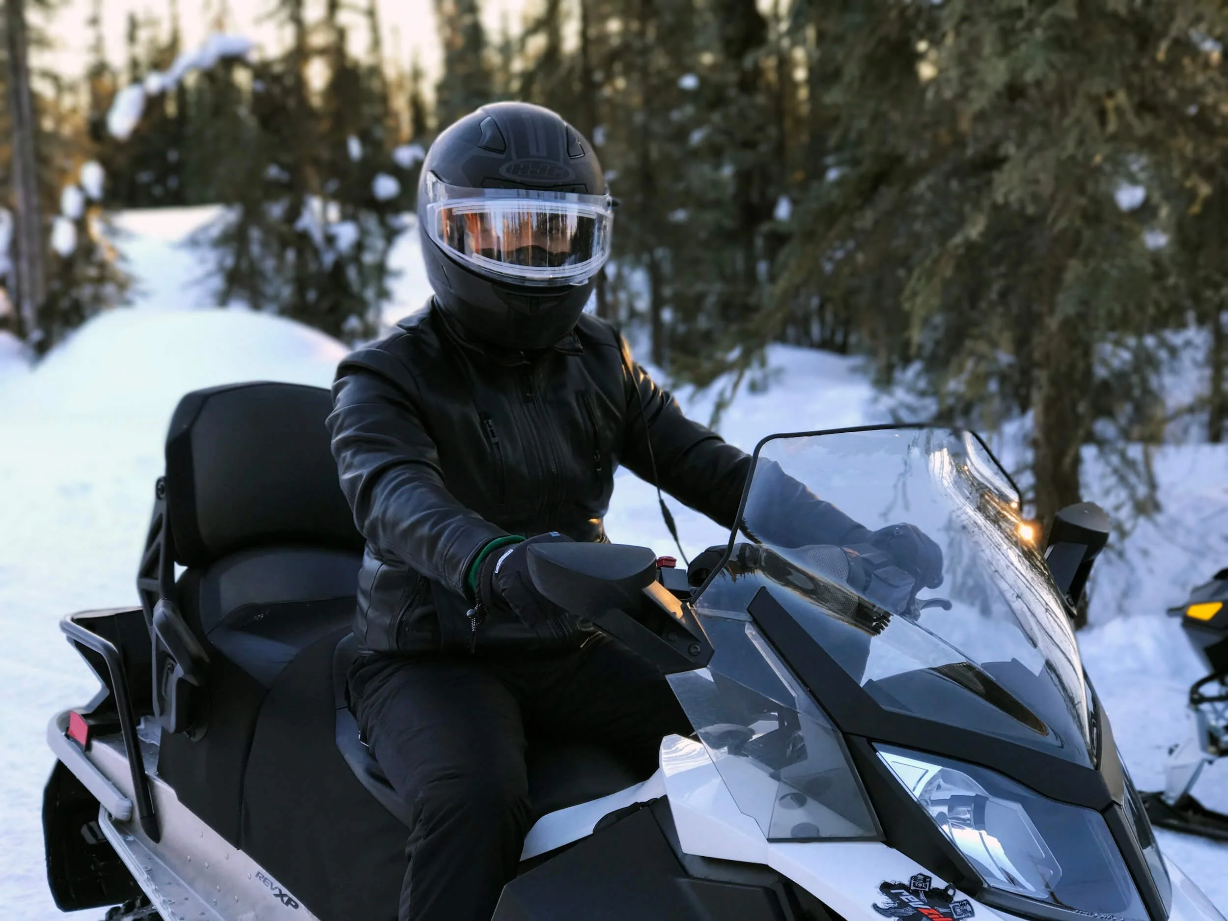 Rider in helmet and black jacket operating white and black snowmobile on snowy forest trail during winter.