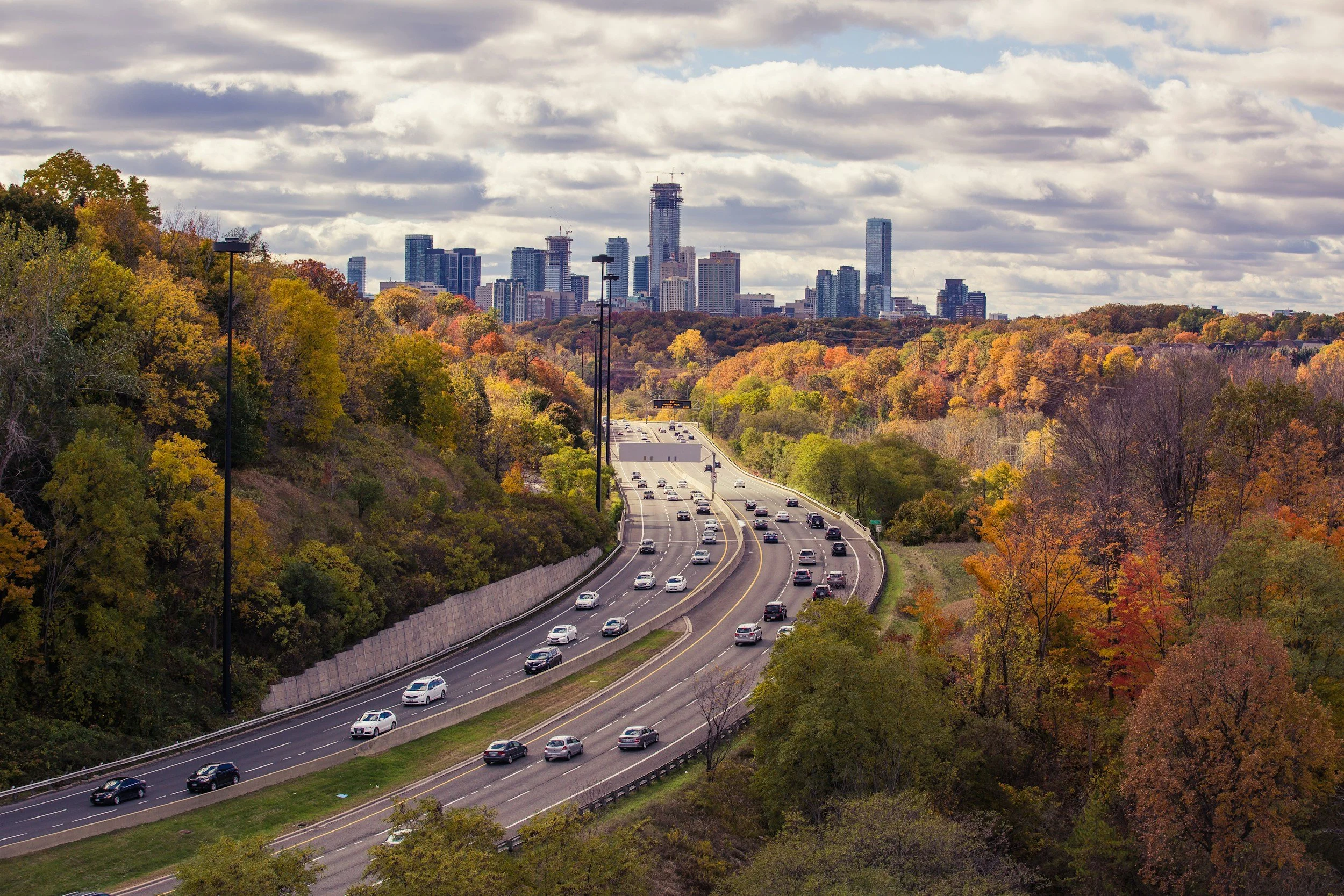 Highway with multiple vehicles driving through forested autumn landscape with city skyline