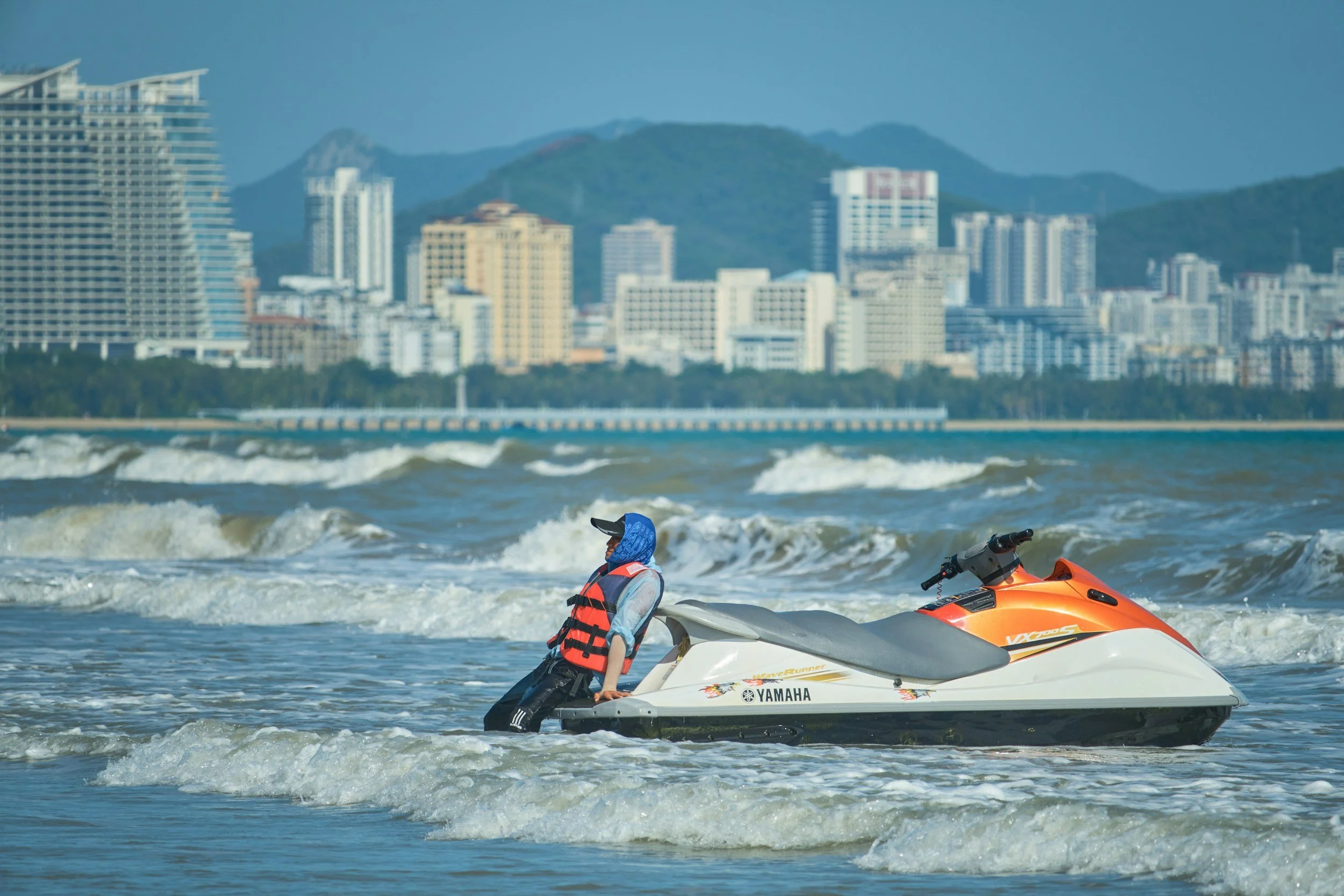 Person riding jet ski in ocean waves with coastal high-rise buildings behind