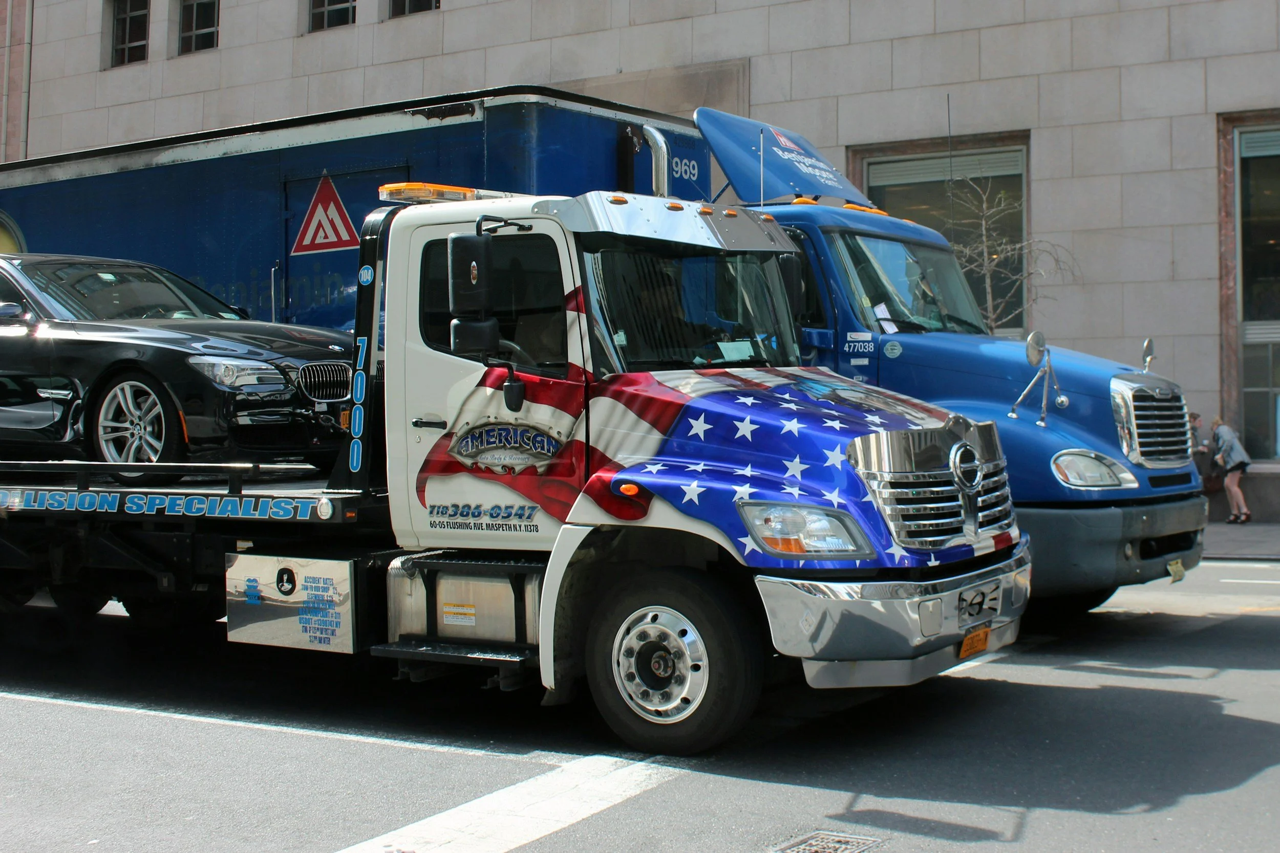 American flag tow truck with collision specialist branding parked on urban street near commercial building