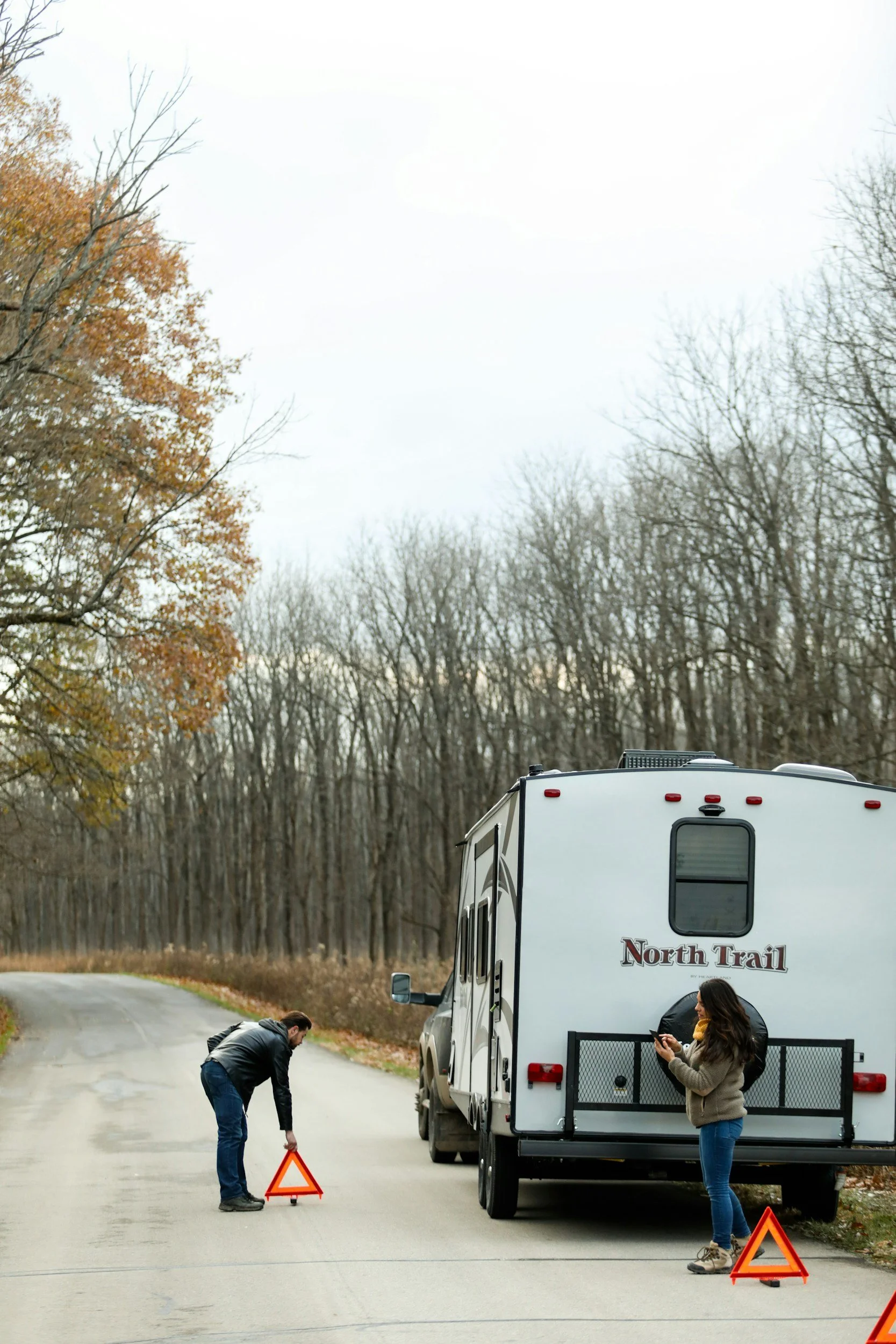 Two people placing warning triangles near a disabled North Trail RV on a rural road with bare trees
