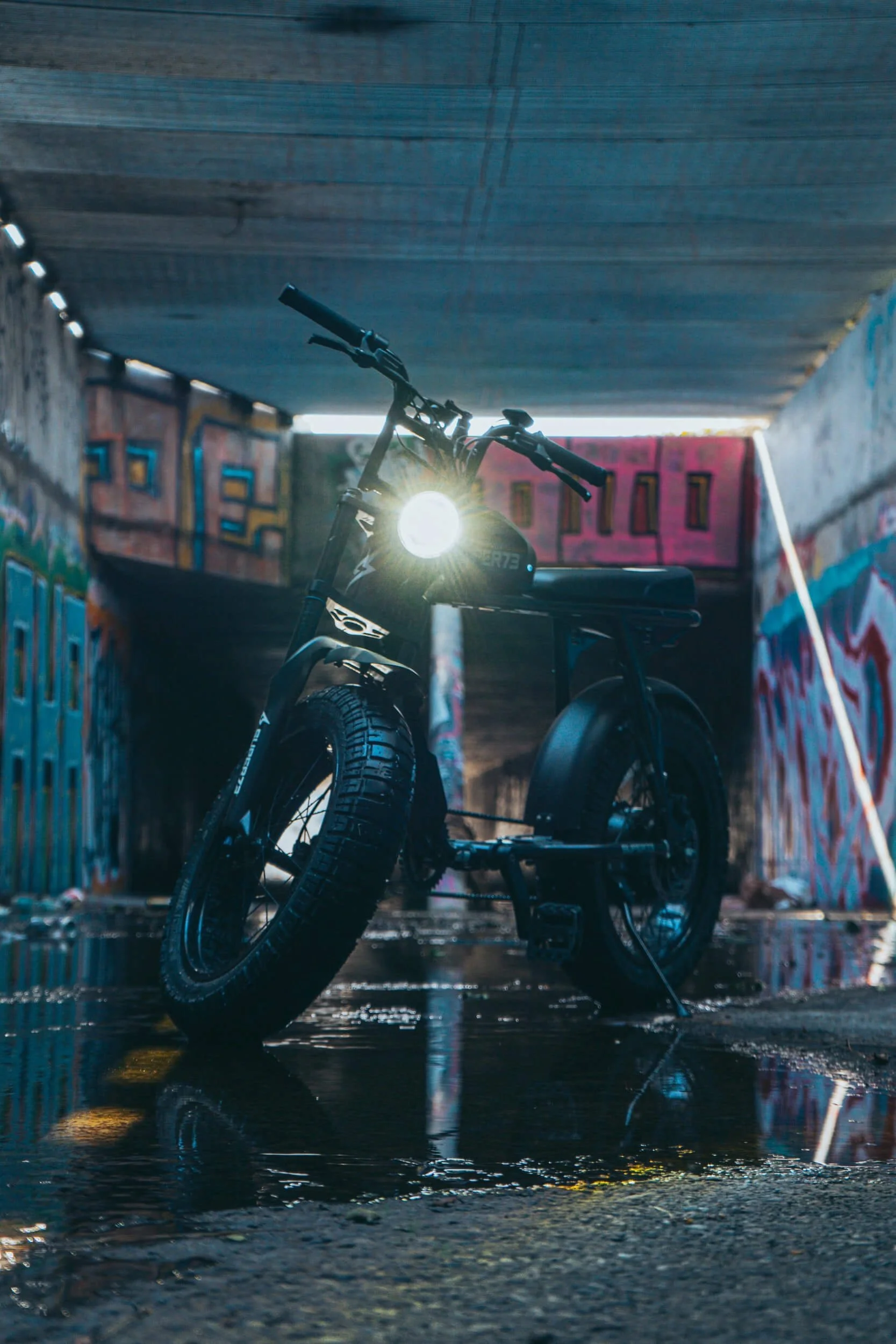 Black electric bike with headlight illuminated in urban alley with colorful graffiti walls and wet pavement