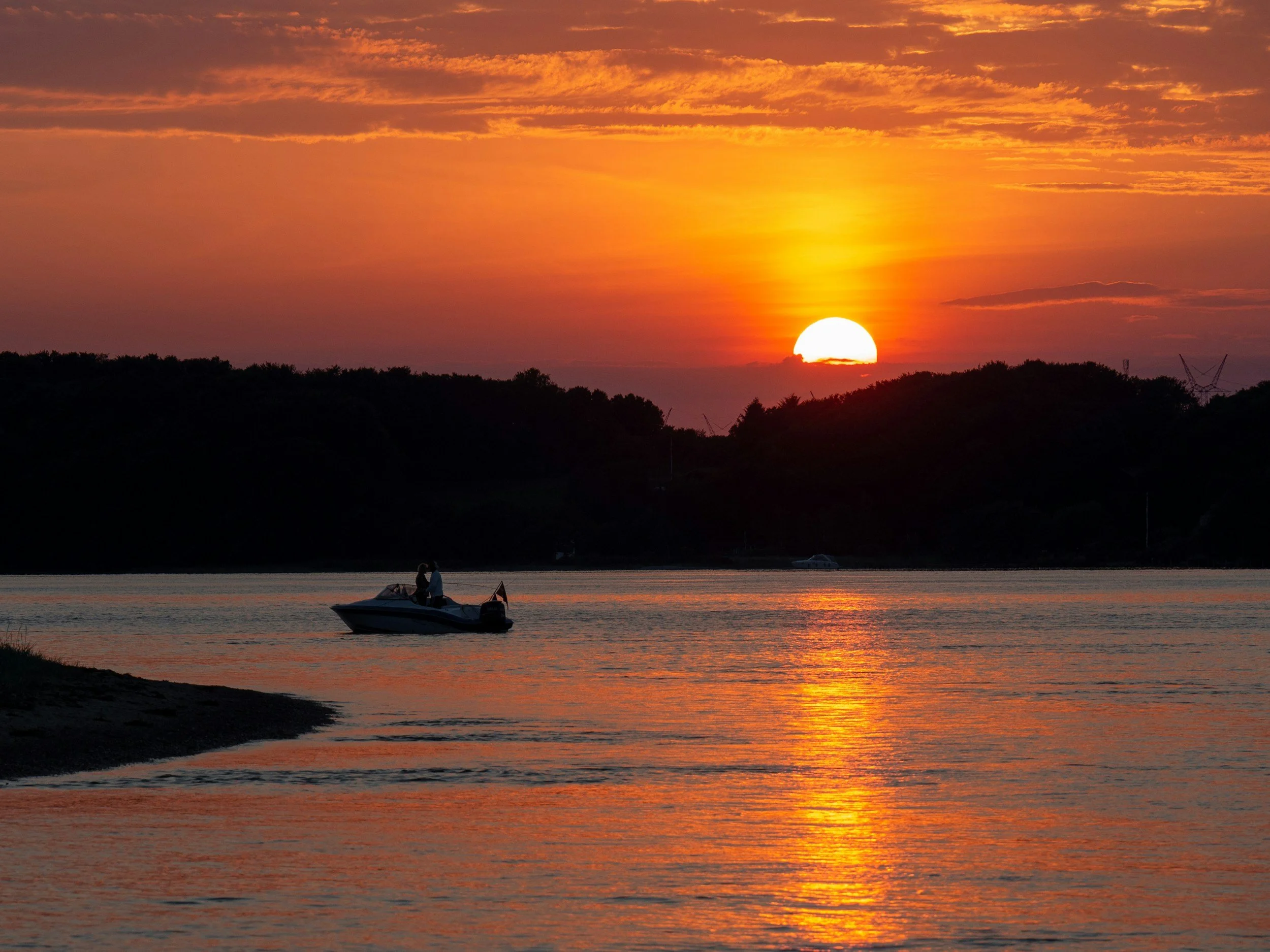 Fishing boat on calm water at sunset with forested shoreline silhouetted against orange sky