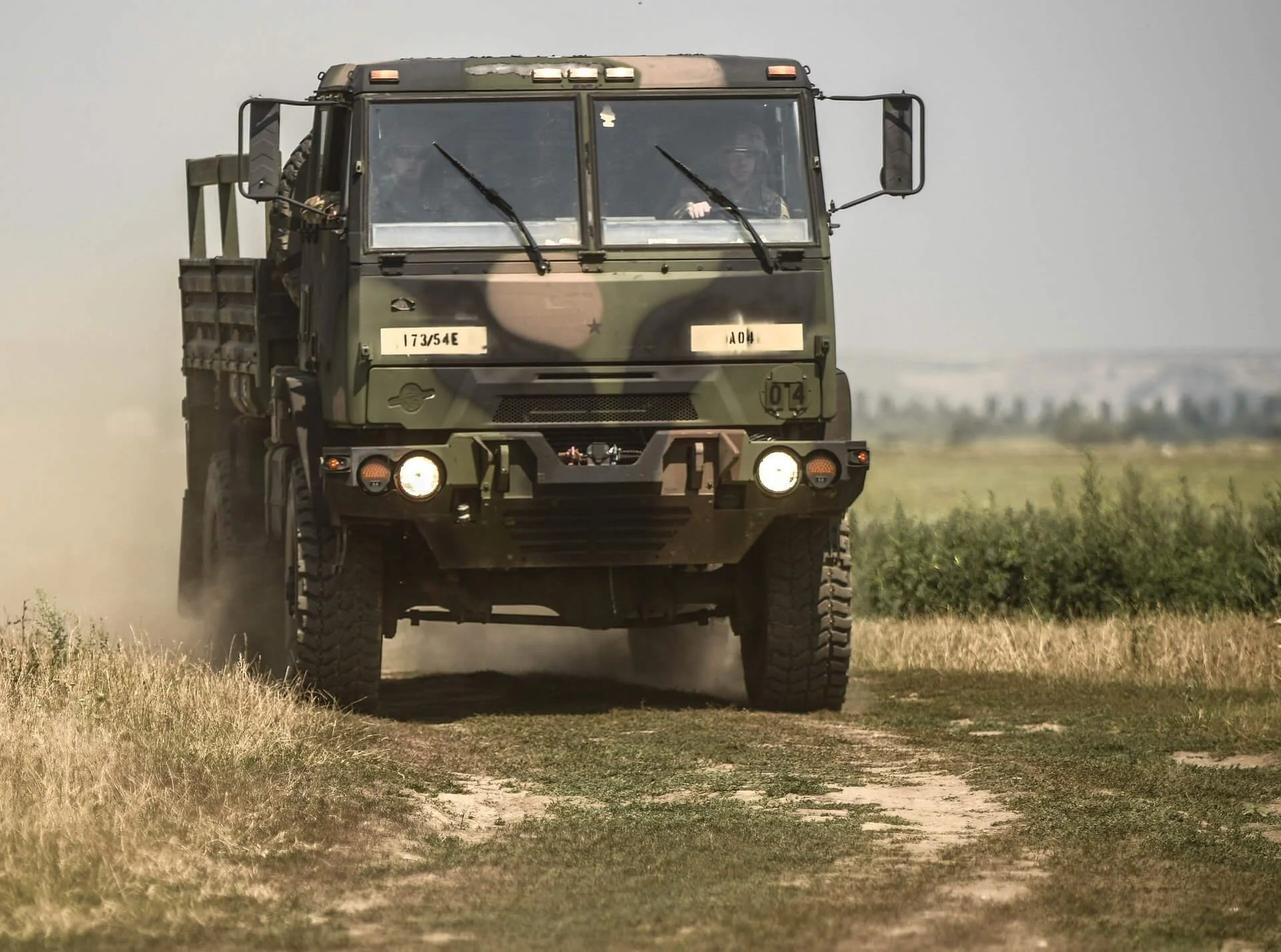 Military LMTV tactical truck driving through dusty rural terrain with cargo bed and armed personnel visible in cab