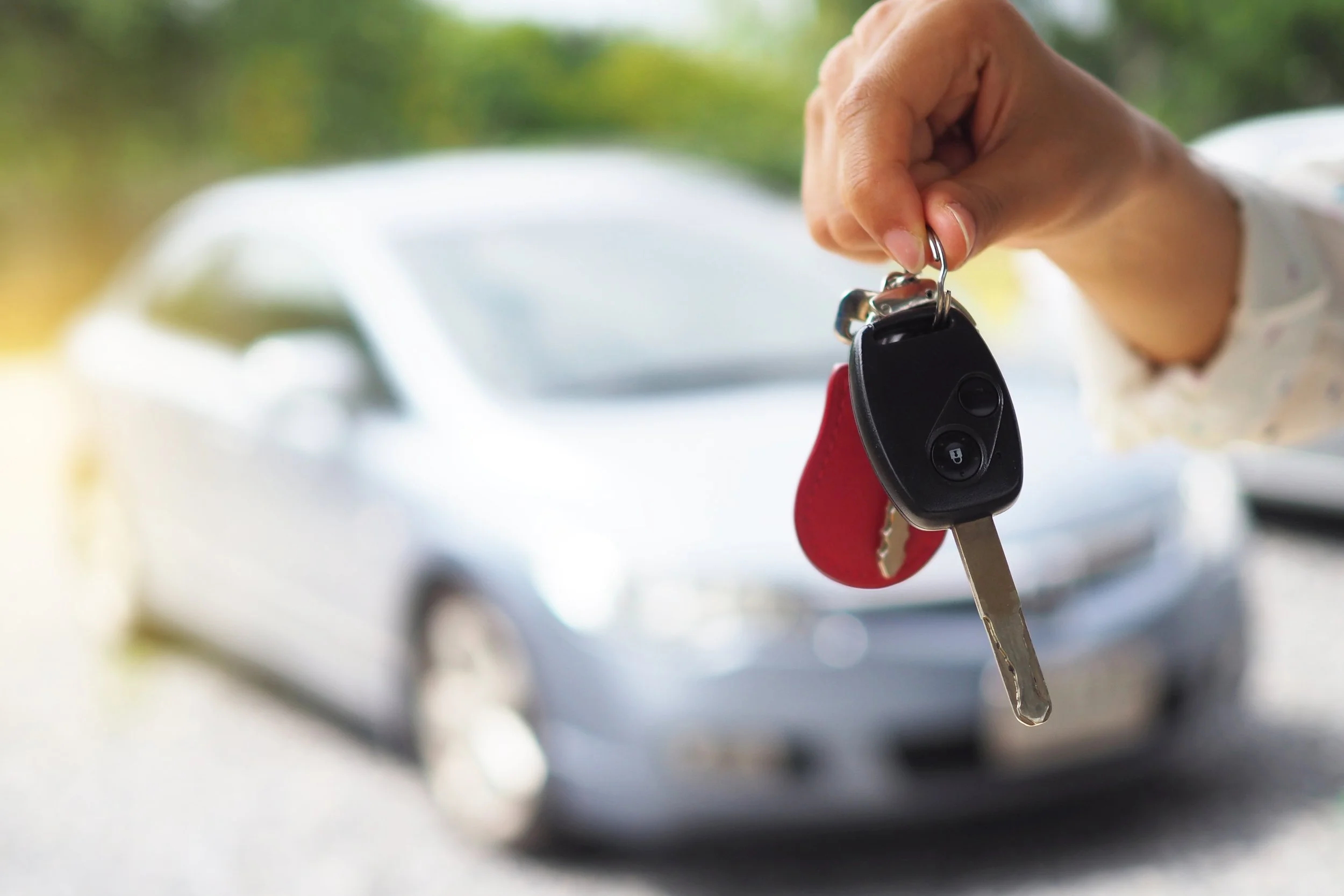 Hand holding car key with red keychain in front of white used vehicle in driveway