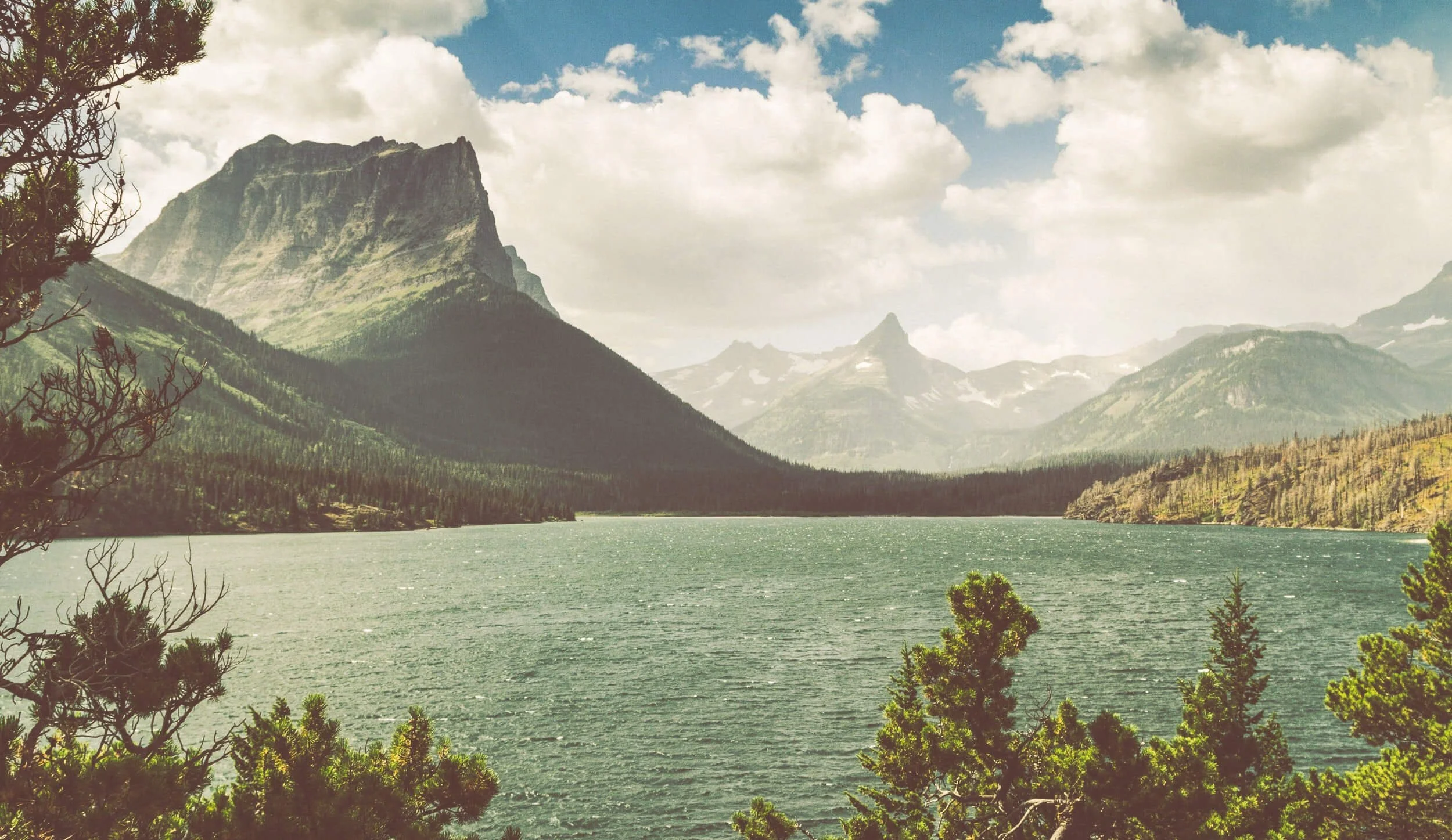 Scenic Montana mountain lake surrounded by evergreen forests and snow-capped peaks under cloudy blue sky