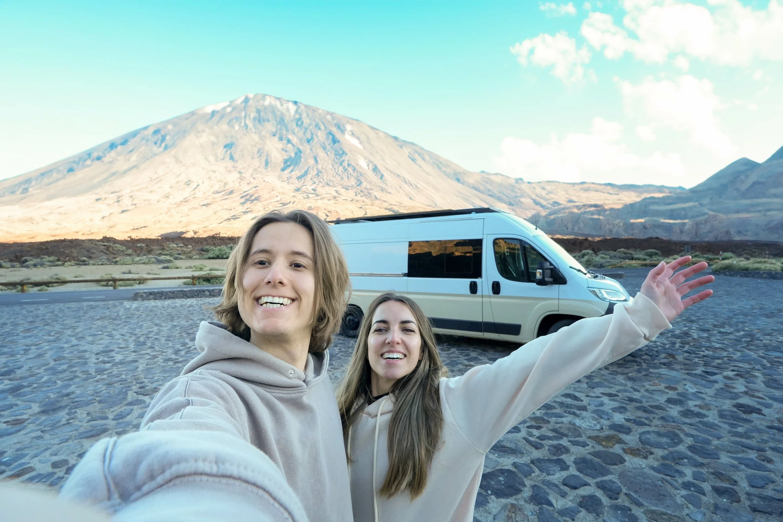Happy couple in white hoodies celebrating RV road trip adventure with camper van and snowy mountain backdrop