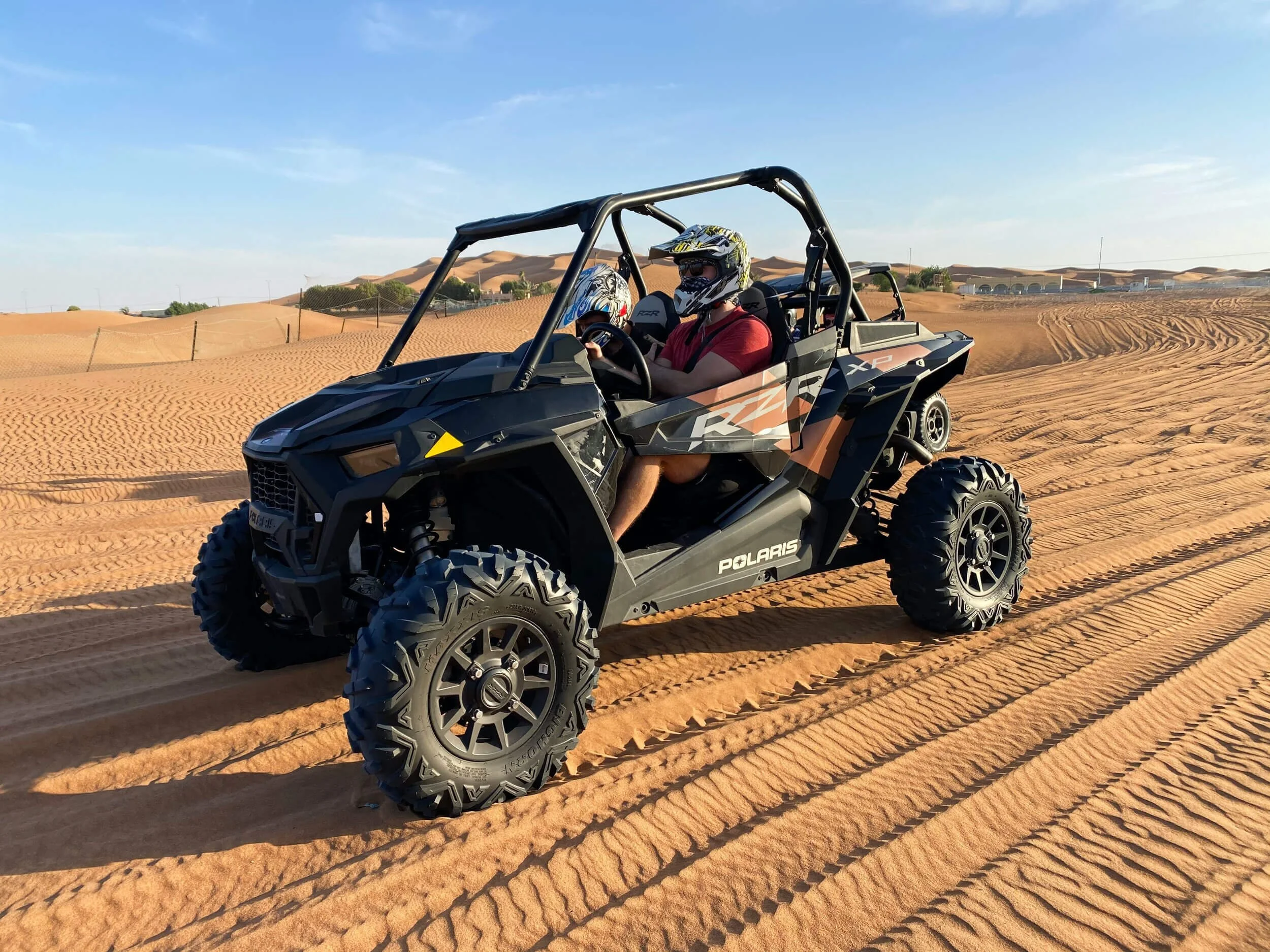Polaris RZR UTV driving on sandy desert terrain with two riders wearing helmets and safety gear