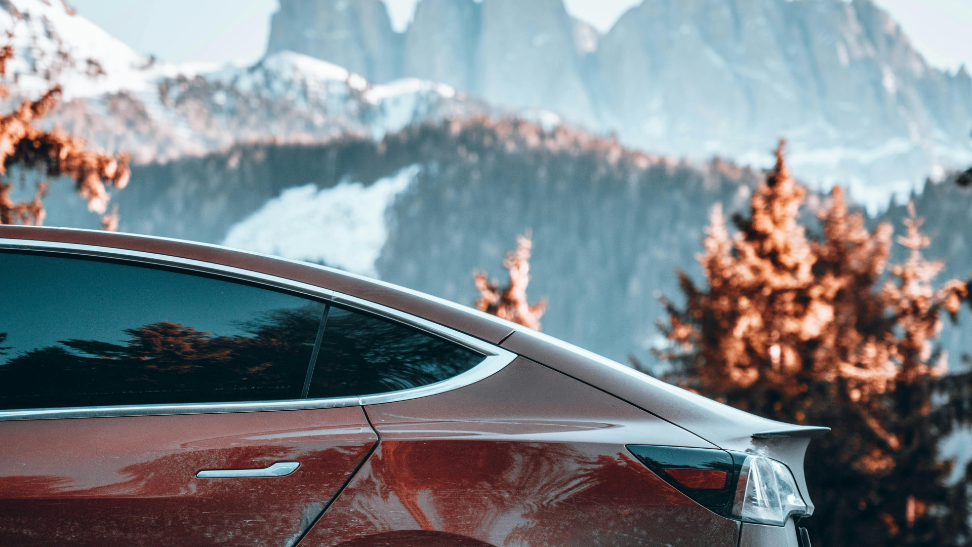 Red SUV parked in snowy mountain landscape with evergreen trees and peaks in background