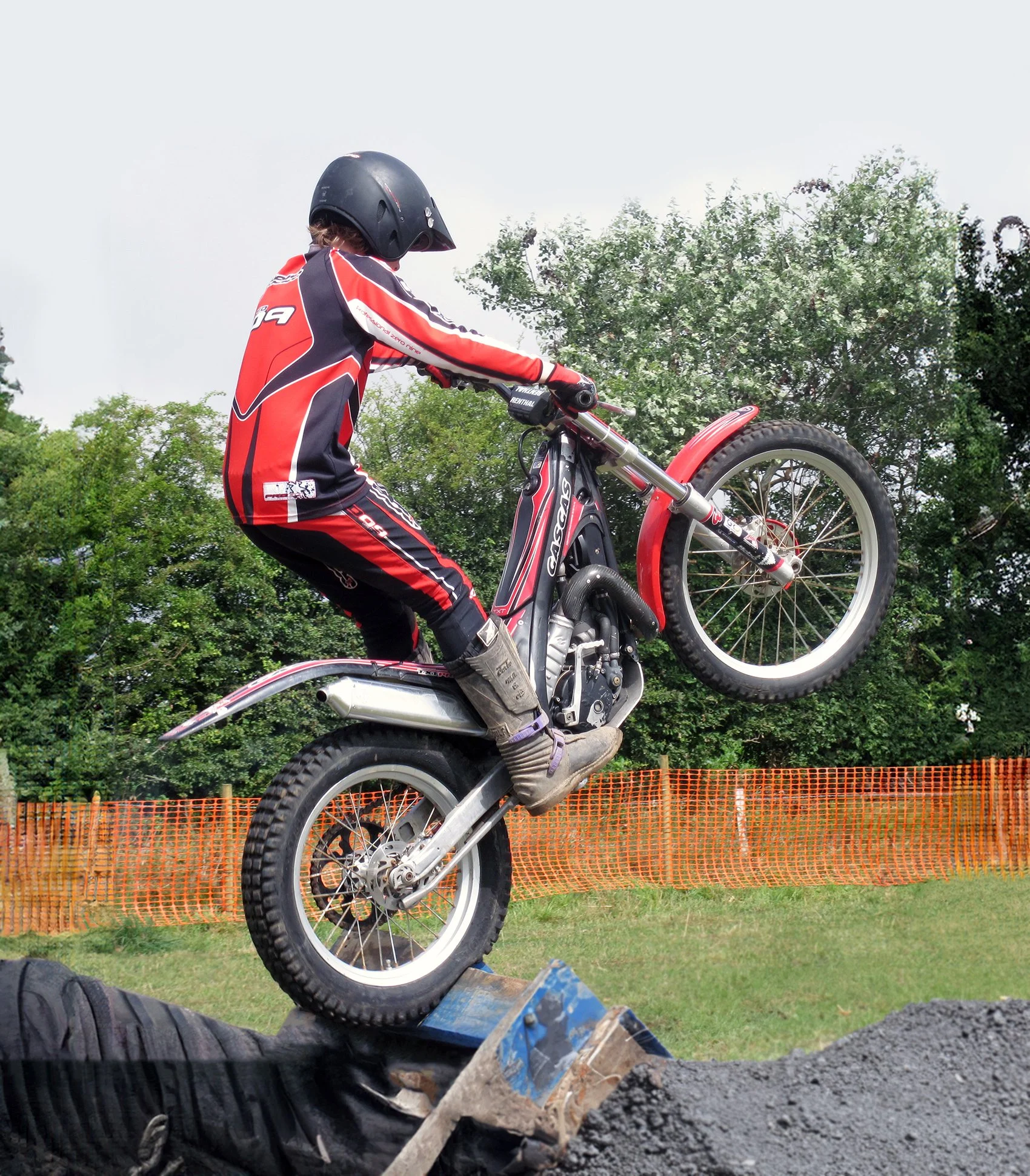 Red trials bike rider performing wheelie stunt over obstacle, demonstrating street-legal motorcycle techniques and skills.
