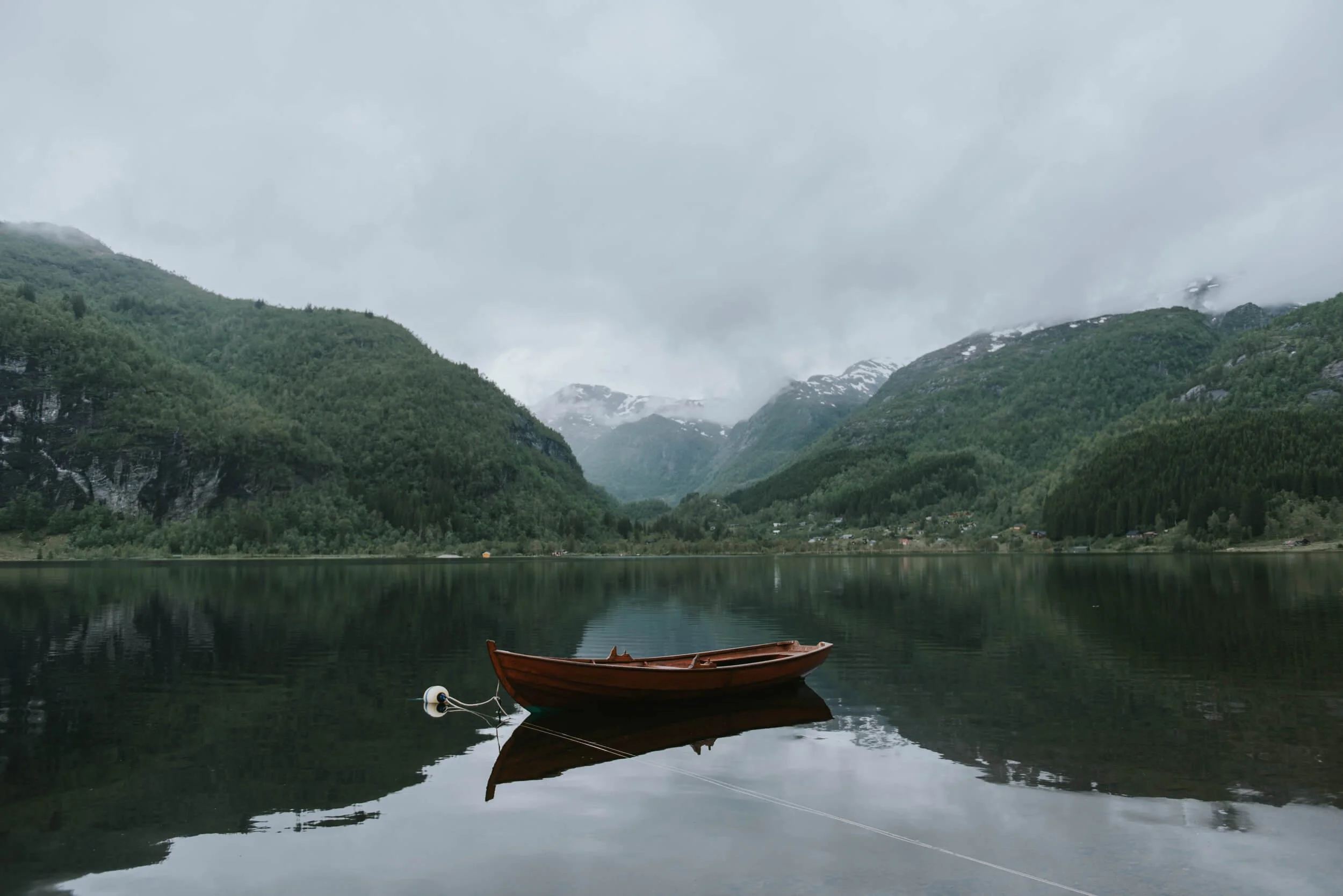 Wooden boat anchored in calm alpine lake surrounded by forested mountains and snow-capped peaks